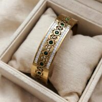 Broad gold kada with central line of oval emerald green stones, jaali filigree cutwork around them, and two side borders of baguette white CZ stones. Single piece heavy kada.